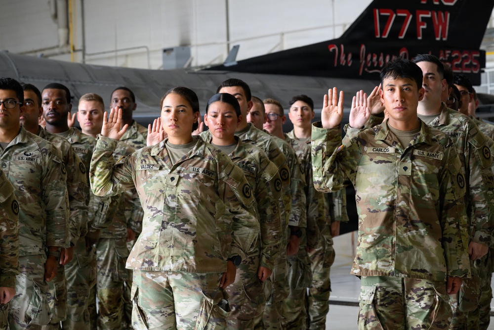 A group of U.S. Army soldiers in OCP (Operational Camouflage Pattern) uniforms are raising their right hands, appearing to take an oath or pledge. The ceremony is taking place inside a military hangar, with a fighter jet bearing '177 FW' (177th Fighter Wing) markings visible in the background. Name tapes visible on soldiers include 'VALDEZ' and 'CARLOS'.