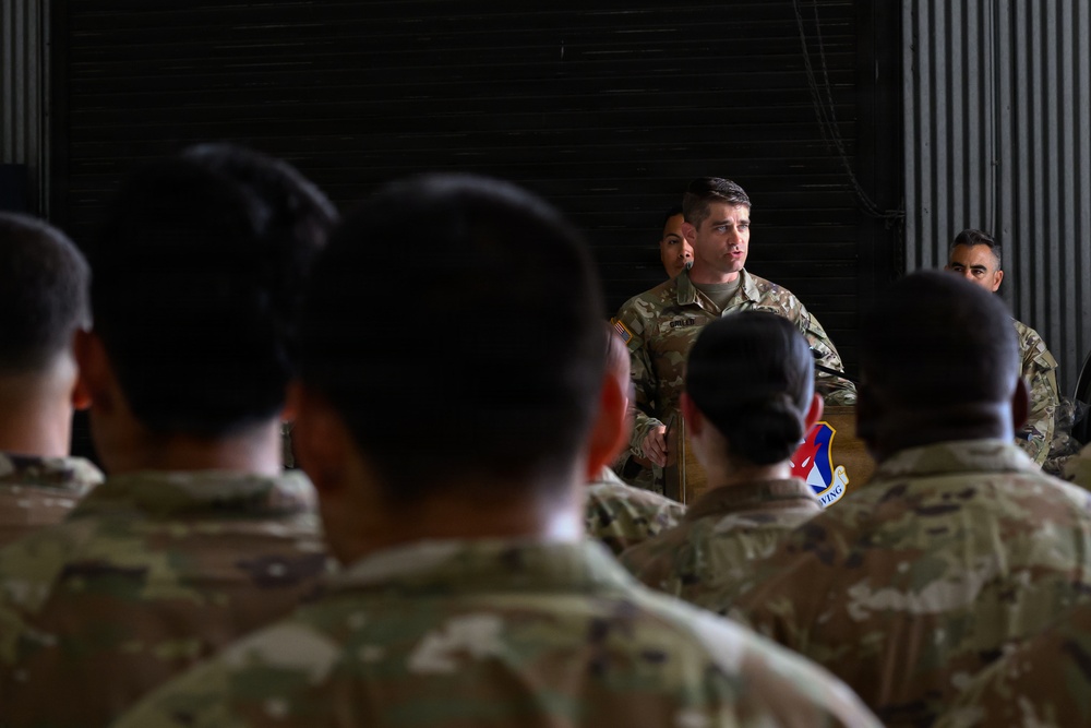 A military officer in camouflage uniform addresses a formation of service members inside what appears to be an aircraft hangar. The speaker stands at a podium bearing a military wing insignia while facing an audience of uniformed personnel with their backs to the camera. The setting and attire suggest a formal military briefing, ceremony, or all-call formation.