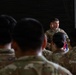 A military officer in camouflage uniform addresses a formation of service members inside what appears to be an aircraft hangar. The speaker stands at a podium bearing a military wing insignia while facing an audience of uniformed personnel with their backs to the camera. The setting and attire suggest a formal military briefing, ceremony, or all-call formation.