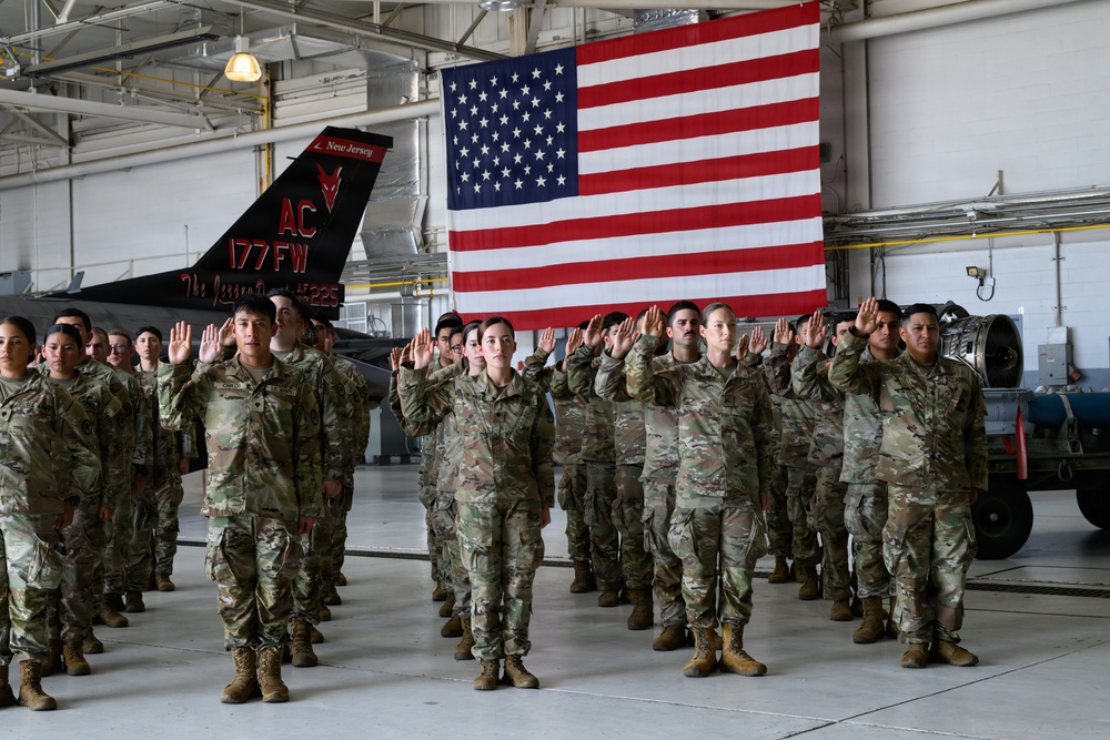 A group of military personnel in camouflage uniforms are raising their right hands in what appears to be a formal oath ceremony inside a military hangar. A large American flag is prominently displayed in the background, and a military aircraft tail fin bearing the markings '177 FW' and 'New Jersey' is visible, indicating this is likely at the 177th Fighter Wing of the New Jersey Air National Guard. The ceremony appears to be a reenlistment or enlistment swearing-in event.