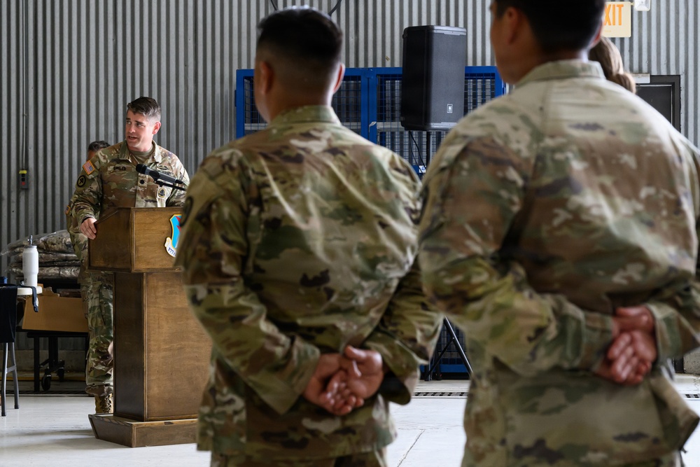 A military ceremony or briefing is taking place inside what appears to be a hangar or large military facility. A soldier in OCP (Operational Camouflage Pattern) uniform is speaking at a wooden podium equipped with a microphone, while two other soldiers stand at parade rest with hands clasped behind their backs. The setting suggests a formal military event such as an awards ceremony, unit briefing, or change of command.