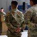 A military ceremony or briefing is taking place inside what appears to be a hangar or large military facility. A soldier in OCP (Operational Camouflage Pattern) uniform is speaking at a wooden podium equipped with a microphone, while two other soldiers stand at parade rest with hands clasped behind their backs. The setting suggests a formal military event such as an awards ceremony, unit briefing, or change of command.