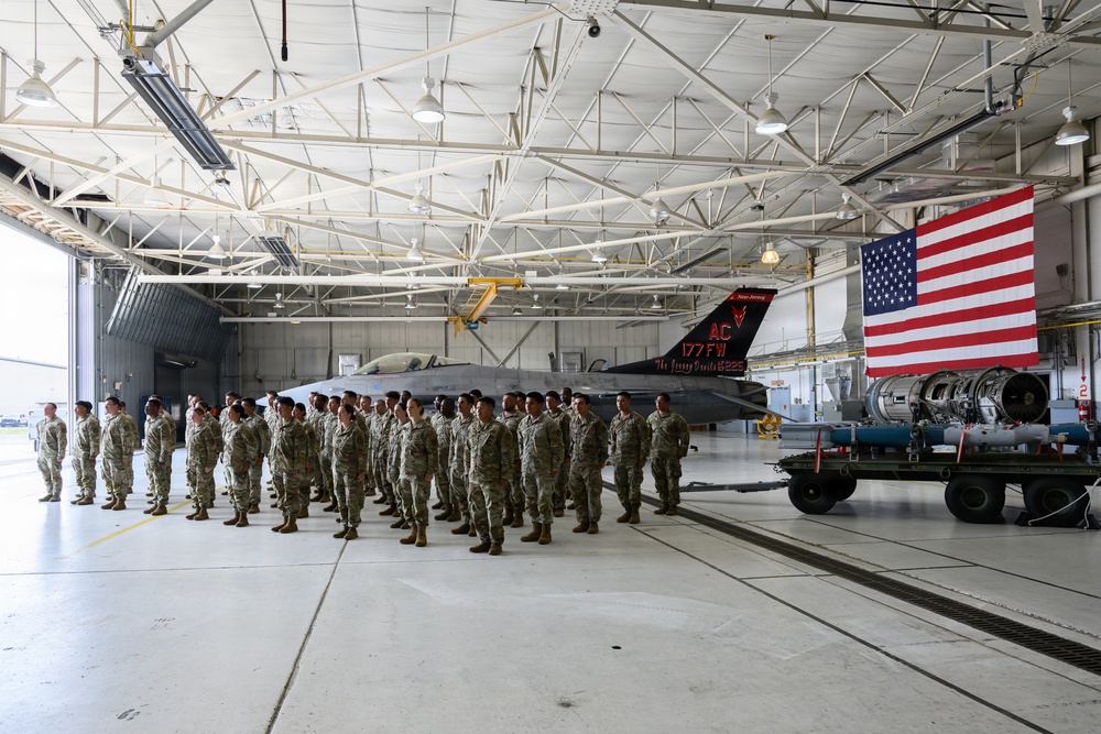 A large formation of military personnel in camouflage uniforms stands at attention inside an aircraft hangar. Behind them is a black-painted F-16 fighter jet bearing the markings 'AC 177FW' and 'New Jersey,' along with the text 'The Jersey Devils.' A large American flag is displayed on the right side of the hangar, suggesting a formal military ceremony or formation.