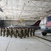 A large formation of military personnel in camouflage uniforms stands at attention inside an aircraft hangar. Behind them is a black-painted F-16 fighter jet bearing the markings 'AC 177FW' and 'New Jersey,' along with the text 'The Jersey Devils.' A large American flag is displayed on the right side of the hangar, suggesting a formal military ceremony or formation.