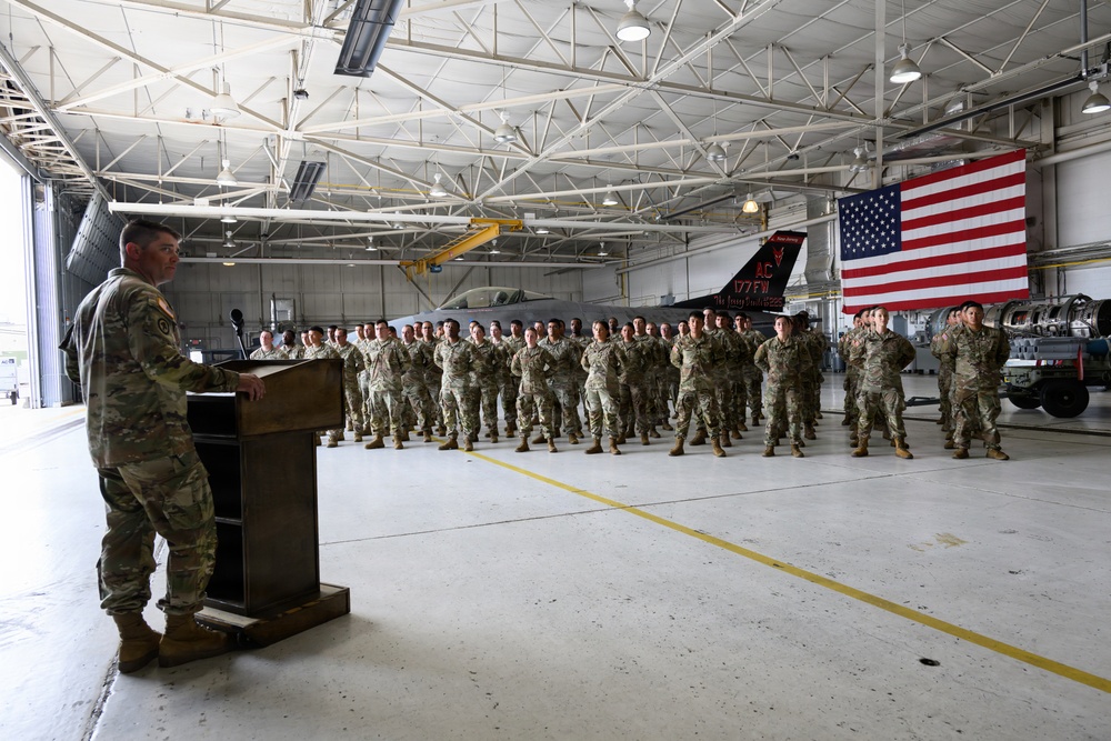 A military officer addresses a large formation of uniformed personnel inside an aircraft hangar. The setting appears to be a formal military ceremony or briefing, with an American flag and a fighter jet visible in the background. The tail markings on the aircraft indicate this is associated with the 177th Fighter Wing.