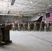 A military officer addresses a large formation of uniformed personnel inside an aircraft hangar. The setting appears to be a formal military ceremony or briefing, with an American flag and a fighter jet visible in the background. The tail markings on the aircraft indicate this is associated with the 177th Fighter Wing.