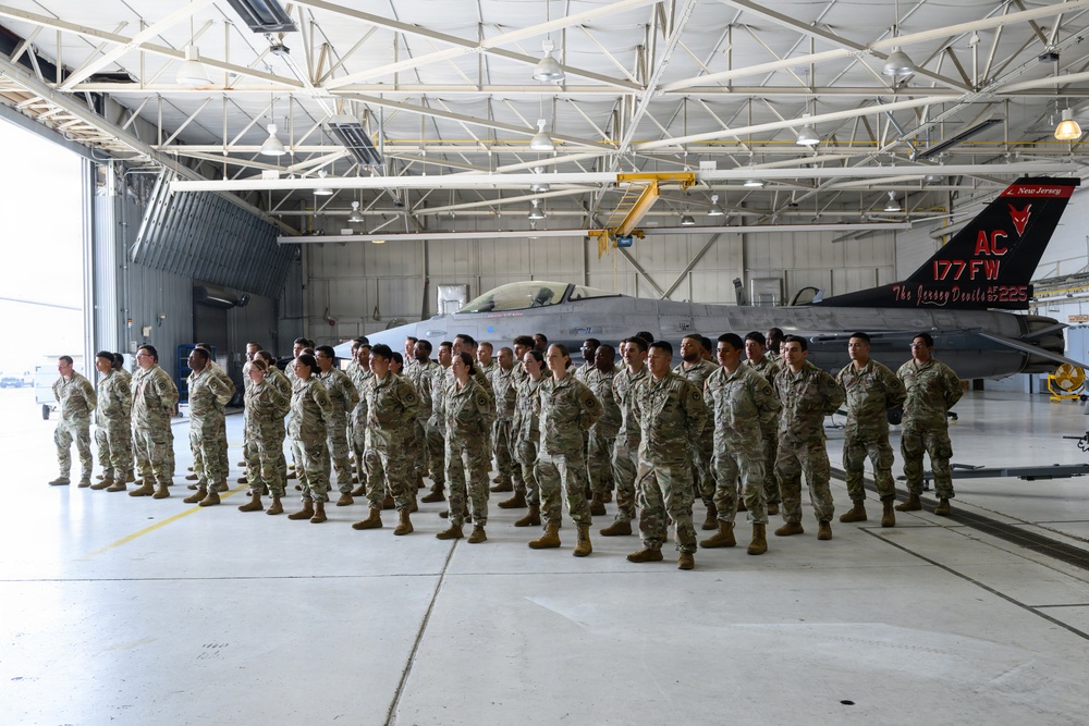 A large group of military personnel in camouflage uniforms are assembled in formation inside an aircraft hangar. Behind them is an F-16 Fighting Falcon jet bearing the markings of the 177th Fighter Wing (177 FW) from New Jersey Air National Guard, with 'AC' tail code and 'The Jersey Devils' nickname. The setting appears to be an official military ceremony or formation event.