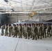 A large group of military personnel in camouflage uniforms are assembled in formation inside an aircraft hangar. Behind them is an F-16 Fighting Falcon jet bearing the markings of the 177th Fighter Wing (177 FW) from New Jersey Air National Guard, with 'AC' tail code and 'The Jersey Devils' nickname. The setting appears to be an official military ceremony or formation event.