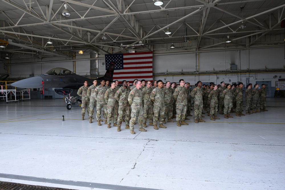 A large formation of military personnel in OCP (Operational Camouflage Pattern) uniforms stands at attention inside a military aircraft hangar. A large American flag is prominently displayed on the back wall, and an F-16 Fighting Falcon jet is visible to the left. The setting suggests a formal military ceremony, parade, or unit formation event.