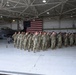 A large formation of military personnel in OCP (Operational Camouflage Pattern) uniforms stands at attention inside a military aircraft hangar. A large American flag is prominently displayed on the back wall, and an F-16 Fighting Falcon jet is visible to the left. The setting suggests a formal military ceremony, parade, or unit formation event.