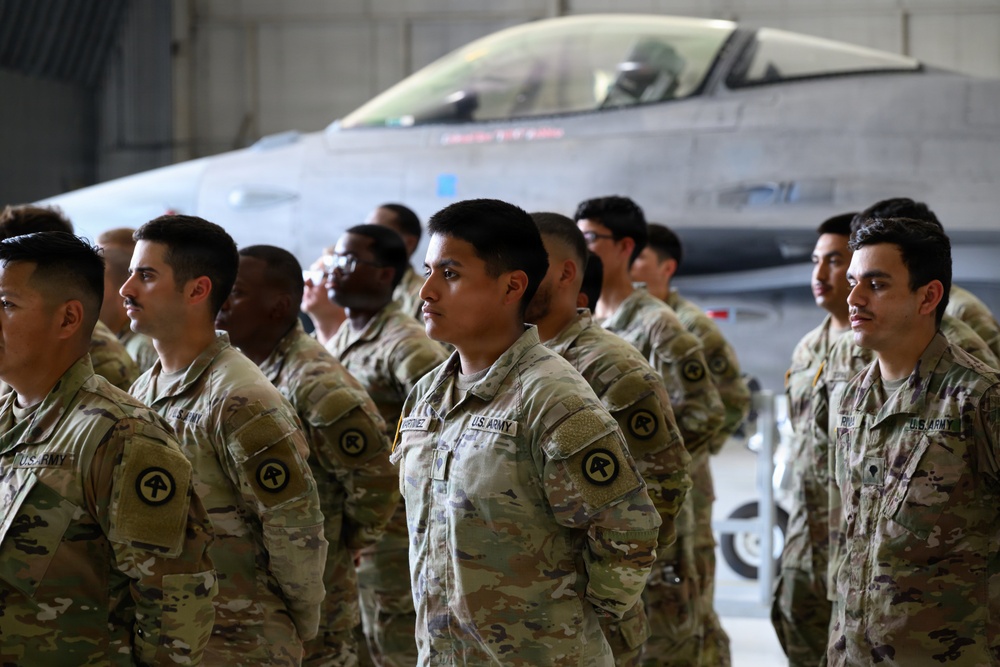 A formation of U.S. Army soldiers in OCP (Operational Camouflage Pattern) uniforms standing at attention inside a military hangar. A fighter jet, appearing to be an F-16, is visible in the background. The soldiers wear shoulder patches consistent with the 4th Infantry Division or similar unit insignia.
