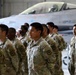 A formation of U.S. Army soldiers in OCP (Operational Camouflage Pattern) uniforms standing at attention inside a military hangar. A fighter jet, appearing to be an F-16, is visible in the background. The soldiers wear shoulder patches consistent with the 4th Infantry Division or similar unit insignia.