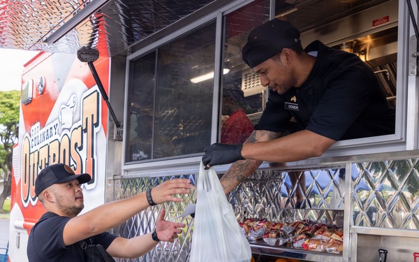 A food truck worker named 'Coger' is handing a plastic bag of food to a customer from 'The Culinary Outpost' food truck. The worker is leaning out of the service window wearing black gloves and a black uniform, while the customer reaches up to receive the order. The truck displays fresh fruit including green apples and oranges, as well as packaged snacks in a lower display shelf.