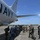 A group of military personnel from multiple nations are gathered on an airport tarmac beside a U.S. Navy aircraft, likely a P-8 Poseidon maritime patrol plane. The personnel are wearing various uniforms indicating a multinational military exercise or cooperative engagement. The scene appears to be a walkthrough, briefing, or tour of the aircraft.