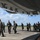 Military personnel from what appears to be multiple nations are gathered on an airfield tarmac beneath the fuselage of a large aircraft. The group includes individuals in flight suits and military uniforms, suggesting a joint or multinational aviation exercise or briefing. The setting appears to be a tropical or subtropical airbase based on the vegetation and cloud formations visible in the background.