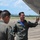 Military personnel in flight suits are gathered on an airfield tarmac next to a white aircraft. A man with a mustache, wearing a green flight suit with patches, appears to be briefing or explaining something about the aircraft to two other personnel. The setting appears to be a tropical or subtropical location based on the vegetation and mountains visible in the background.