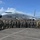 A large group of military personnel from multiple nations pose for a group photograph in front of a U.S. Navy P-8A Poseidon maritime patrol aircraft bearing the number '335' on the tarmac. Personnel are wearing a variety of military uniforms including flight suits, Navy working uniforms, and camouflage patterns, suggesting a multinational military exercise or cooperative event. The setting appears to be a tropical airfield, indicated by the lush green hills and vegetation visible in the background.