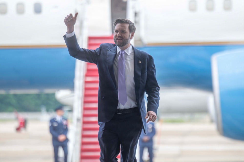 A man in a dark navy suit with a lavender tie is walking on an airport tarmac, giving a thumbs-up gesture toward the camera. Behind him is a large aircraft with red boarding stairs, consistent with official government air travel. Military or official personnel in uniform are visible in the blurred background.