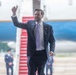 A man in a dark navy suit with a lavender tie is walking on an airport tarmac, giving a thumbs-up gesture toward the camera. Behind him is a large aircraft with red boarding stairs, consistent with official government air travel. Military or official personnel in uniform are visible in the blurred background.