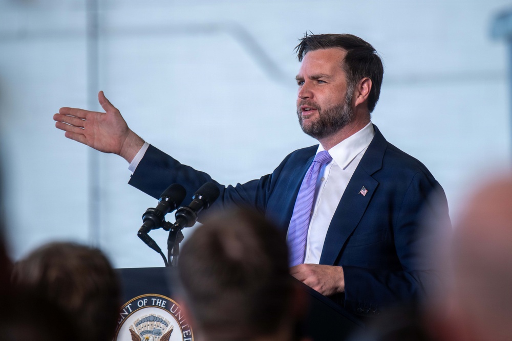 A man in a dark navy suit with a light purple tie is speaking at a podium equipped with microphones. He is gesturing with his right hand while addressing an audience. A partial seal reading 'ent of the Unite...' is visible on the podium, suggesting an official government setting.