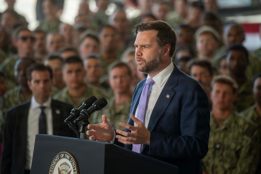 A man in a dark navy suit with a lavender tie is speaking at a podium bearing a partial seal that reads 'President of the Unite[d States]'. He is gesturing with both hands while addressing a large crowd of individuals dressed in military camouflage uniforms standing behind him. The setting appears to be a military hangar or large indoor facility.