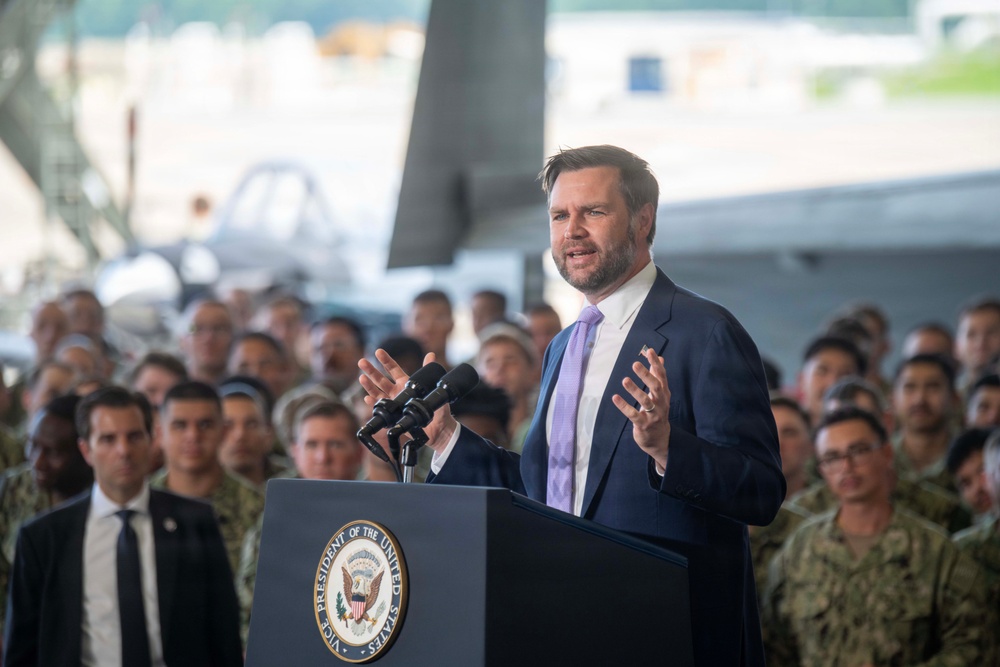 A man in a navy blue suit and lavender tie is speaking at a podium bearing the seal of the Vice President of the United States. He is addressing a large crowd of what appear to be military personnel in uniform, gathered inside what looks like a military hangar. Military aircraft are partially visible in the background.