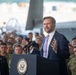 A man in a navy blue suit and lavender tie is speaking at a podium bearing the seal of the Vice President of the United States. He is addressing a large crowd of what appear to be military personnel in uniform, gathered inside what looks like a military hangar. Military aircraft are partially visible in the background.