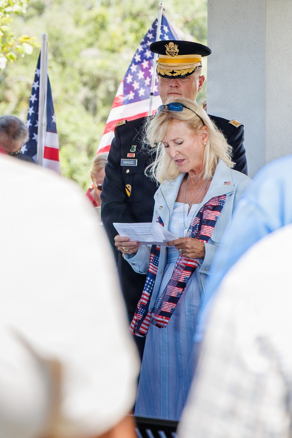 A patriotic ceremony, likely a Memorial Day or Veterans Day event, is taking place outdoors. A blonde woman in a light blue jacket with an American flag scarf is reading from a paper, while a U.S. Army officer in dress uniform stands behind her. Multiple American flags are visible in the background.