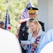 A patriotic ceremony, likely a Memorial Day or Veterans Day event, is taking place outdoors. A blonde woman in a light blue jacket with an American flag scarf is reading from a paper, while a U.S. Army officer in dress uniform stands behind her. Multiple American flags are visible in the background.
