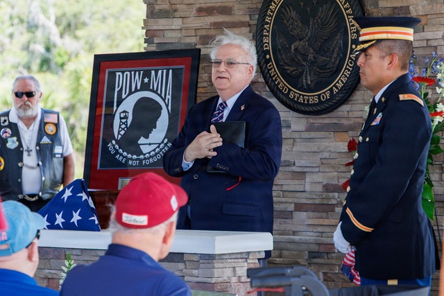 A formal military/veterans ceremony is taking place at what appears to be a Department of Veterans Affairs facility in the United States. A civilian speaker in a navy suit is addressing an audience in front of a framed POW/MIA flag displaying the text 'You Are Not Forgotten.' A uniformed U.S. Army officer stands at attention to the right, while a veteran in a biker vest observes from the left.
