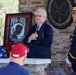 A formal military/veterans ceremony is taking place at what appears to be a Department of Veterans Affairs facility in the United States. A civilian speaker in a navy suit is addressing an audience in front of a framed POW/MIA flag displaying the text 'You Are Not Forgotten.' A uniformed U.S. Army officer stands at attention to the right, while a veteran in a biker vest observes from the left.