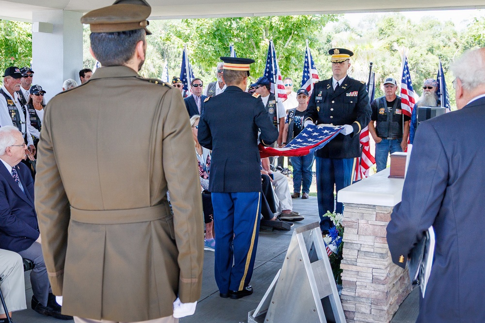 A military funeral or memorial ceremony is taking place outdoors under a covered pavilion. Two U.S. Army soldiers in dress uniforms are performing a flag-folding ceremony over what appears to be a casket or memorial stand, while attendees including veterans, civilians, and other military personnel look on. Multiple American flags and what appear to be POW/MIA flags are visible in the background.