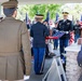 A military funeral or memorial ceremony is taking place outdoors under a covered pavilion. Two U.S. Army soldiers in dress uniforms are performing a flag-folding ceremony over what appears to be a casket or memorial stand, while attendees including veterans, civilians, and other military personnel look on. Multiple American flags and what appear to be POW/MIA flags are visible in the background.