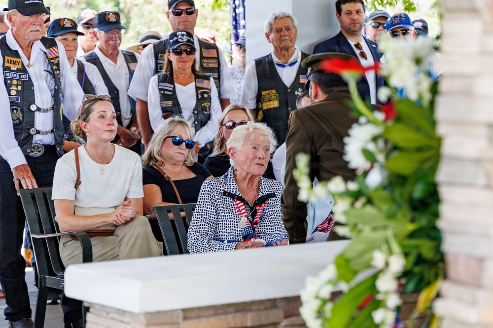 A solemn military or veterans memorial ceremony is taking place outdoors, with attendees seated and standing in observation. Several individuals wearing veteran motorcycle club vests with patches are visible in the background, while civilians, including an elderly woman holding small American flags, are seated in the foreground. A floral wreath is prominently displayed in the right foreground, suggesting a commemorative or burial service.