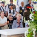 A solemn military or veterans memorial ceremony is taking place outdoors, with attendees seated and standing in observation. Several individuals wearing veteran motorcycle club vests with patches are visible in the background, while civilians, including an elderly woman holding small American flags, are seated in the foreground. A floral wreath is prominently displayed in the right foreground, suggesting a commemorative or burial service.