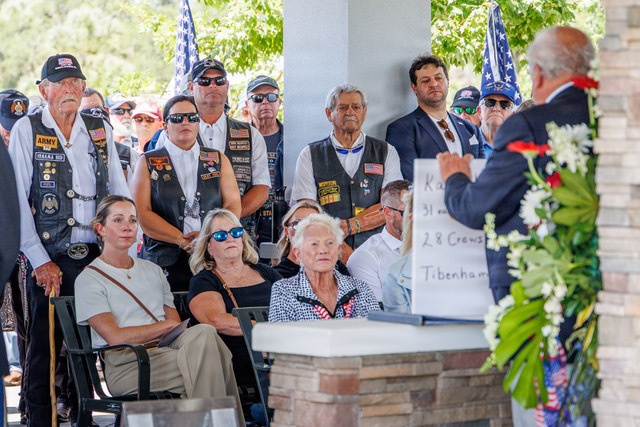 A memorial or commemorative ceremony is taking place outdoors under a covered pavilion. A speaker stands at a podium/lectern decorated with a white floral wreath, addressing an audience that includes veterans wearing motorcycle club vests with military patches and civilians seated in chairs. A whiteboard behind the speaker displays partial text including numbers '31,' '28 Crews,' and 'Tibenham,' suggesting a WWII or military unit commemoration.