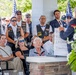 A memorial or commemorative ceremony is taking place outdoors under a covered pavilion. A speaker stands at a podium/lectern decorated with a white floral wreath, addressing an audience that includes veterans wearing motorcycle club vests with military patches and civilians seated in chairs. A whiteboard behind the speaker displays partial text including numbers '31,' '28 Crews,' and 'Tibenham,' suggesting a WWII or military unit commemoration.