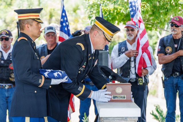 A military funeral or memorial ceremony is taking place outdoors, with two U.S. Army officers in dress uniforms handling a cremation urn and a folded American flag. Veterans and civilians stand in the background, some holding American flags, suggesting this is a formal military burial or interment service. The solemn ceremony appears to honor a deceased veteran.