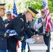 A military funeral or memorial ceremony is taking place outdoors, with two U.S. Army officers in dress uniforms handling a cremation urn and a folded American flag. Veterans and civilians stand in the background, some holding American flags, suggesting this is a formal military burial or interment service. The solemn ceremony appears to honor a deceased veteran.