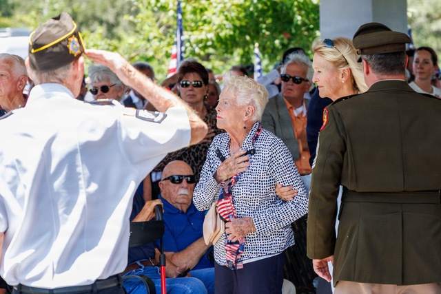 A patriotic ceremony, likely a Memorial Day or Veterans Day event, is taking place outdoors. A veteran in a white uniform and military cap is saluting, while an elderly woman places her hand over her heart, and a person in an olive military dress uniform stands nearby. A crowd of attendees, including several older individuals, observes the proceedings in the background.