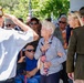 A patriotic ceremony, likely a Memorial Day or Veterans Day event, is taking place outdoors. A veteran in a white uniform and military cap is saluting, while an elderly woman places her hand over her heart, and a person in an olive military dress uniform stands nearby. A crowd of attendees, including several older individuals, observes the proceedings in the background.
