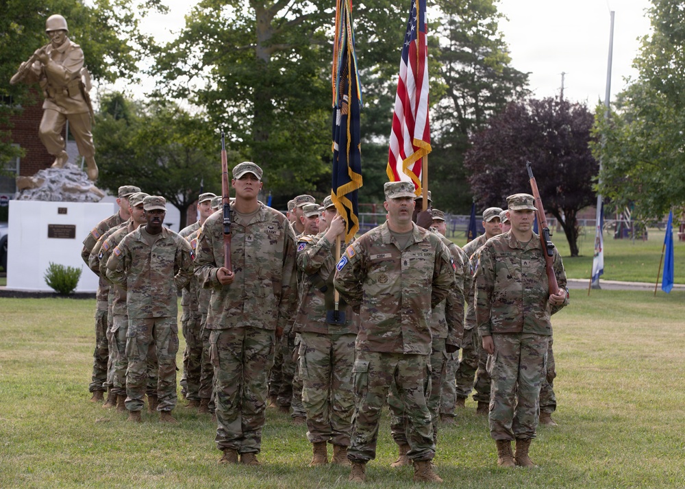 A U.S. Army color guard and formation stand at attention on a grassy parade field during a military ceremony. Soldiers in OCP (Operational Camouflage Pattern) uniforms carry the American flag and what appears to be a unit guidon, flanked by rifle-bearing honor guards. A bronze soldier statue monument is visible in the background left.