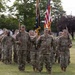 A U.S. Army color guard and formation stand at attention on a grassy parade field during a military ceremony. Soldiers in OCP (Operational Camouflage Pattern) uniforms carry the American flag and what appears to be a unit guidon, flanked by rifle-bearing honor guards. A bronze soldier statue monument is visible in the background left.