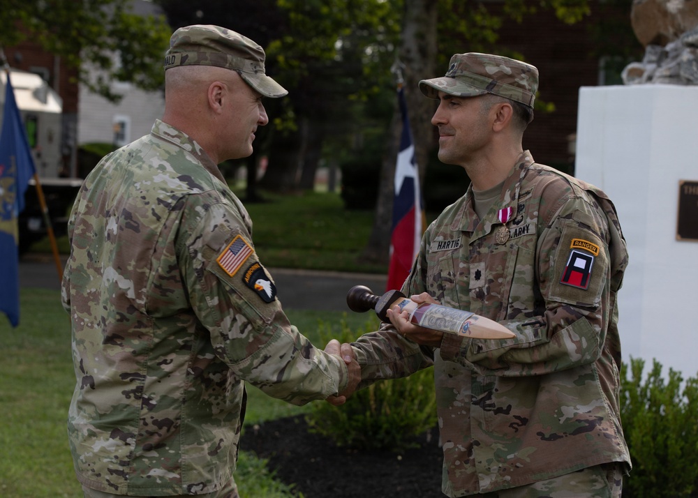 Two U.S. Army soldiers in OCP (Operational Camouflage Pattern) uniforms are shaking hands during what appears to be a military ceremony. The soldier on the right, identified by name tape as 'HARTIG,' is receiving or exchanging a ceremonial item resembling a gavel or mallet. The event appears to be an outdoor change of command or award ceremony.