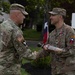 Two U.S. Army soldiers in OCP (Operational Camouflage Pattern) uniforms are shaking hands during what appears to be a military ceremony. The soldier on the right, identified by name tape as 'HARTIG,' is receiving or exchanging a ceremonial item resembling a gavel or mallet. The event appears to be an outdoor change of command or award ceremony.