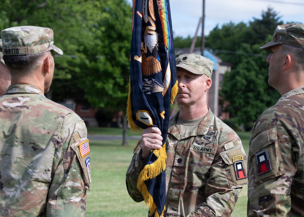 A U.S. Army change of command or unit ceremony is taking place outdoors on a grassy parade field. A lieutenant colonel (identified by the oak leaf insignia on his chest) is holding a unit guidon or colors with gold fringe, a central element of formal military ceremonies. Multiple soldiers in OCP (Operational Camouflage Pattern) uniforms are present, with visible unit patches and tabs.