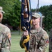 A U.S. Army change of command or unit ceremony is taking place outdoors on a grassy parade field. A lieutenant colonel (identified by the oak leaf insignia on his chest) is holding a unit guidon or colors with gold fringe, a central element of formal military ceremonies. Multiple soldiers in OCP (Operational Camouflage Pattern) uniforms are present, with visible unit patches and tabs.