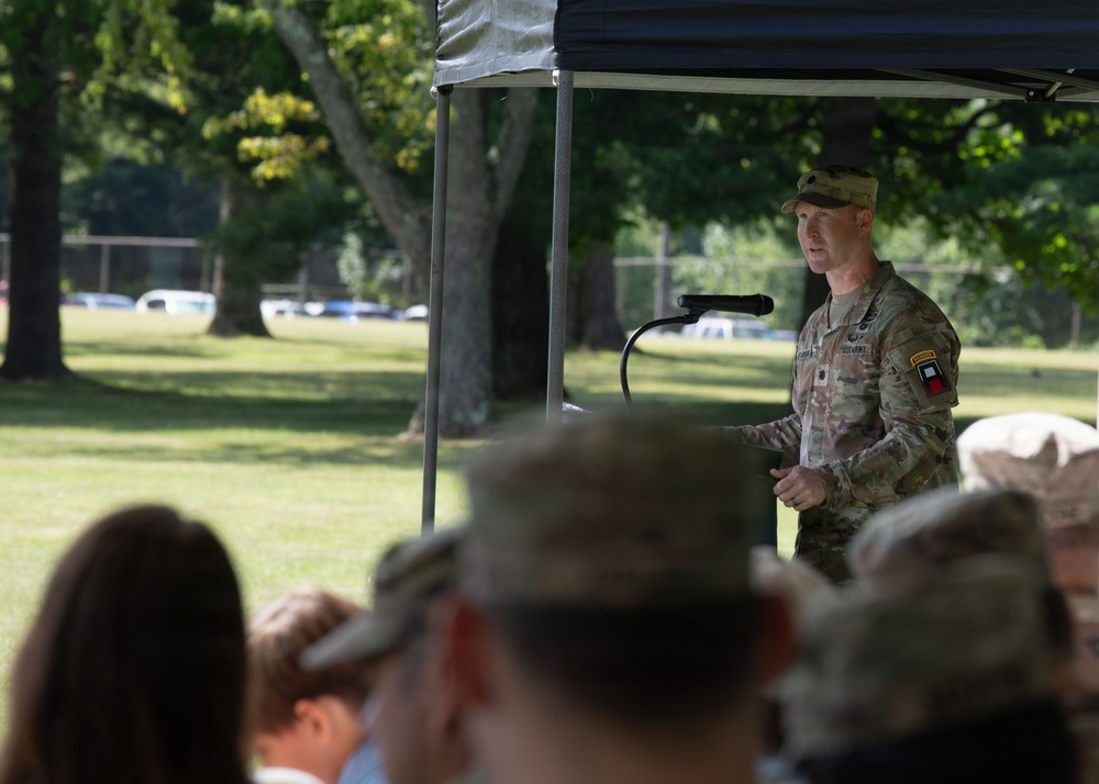 A U.S. Army soldier in Operational Camouflage Pattern (OCP) uniform is speaking at a podium microphone during an outdoor military ceremony or event. The event appears to be held under a canopy tent on a grassy area, with an audience seated in the foreground. Trees and parked vehicles are visible in the background.