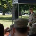 A U.S. Army soldier in Operational Camouflage Pattern (OCP) uniform is speaking at a podium microphone during an outdoor military ceremony or event. The event appears to be held under a canopy tent on a grassy area, with an audience seated in the foreground. Trees and parked vehicles are visible in the background.