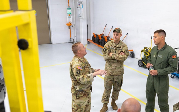 Five military personnel are gathered in what appears to be a hangar or maintenance facility, engaged in a briefing or discussion. One individual in US Army camouflage uniform appears to be explaining something, while others listen, including one person in green flight suit and two individuals wearing patches consistent with a foreign military. The setting features yellow industrial equipment, a polished white floor with yellow safety markings, and maintenance tools in the background.
